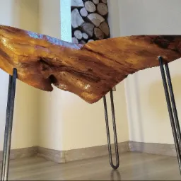 Stolik kawowy z blatem z nieregularnego kawałka drewna o ciepłym, bursztynowym odcieniu, wsparty na metalowych nogach typu hairpin, z widocznymi słojami i naturalnymi pęknięciami drewna, ustawiony...