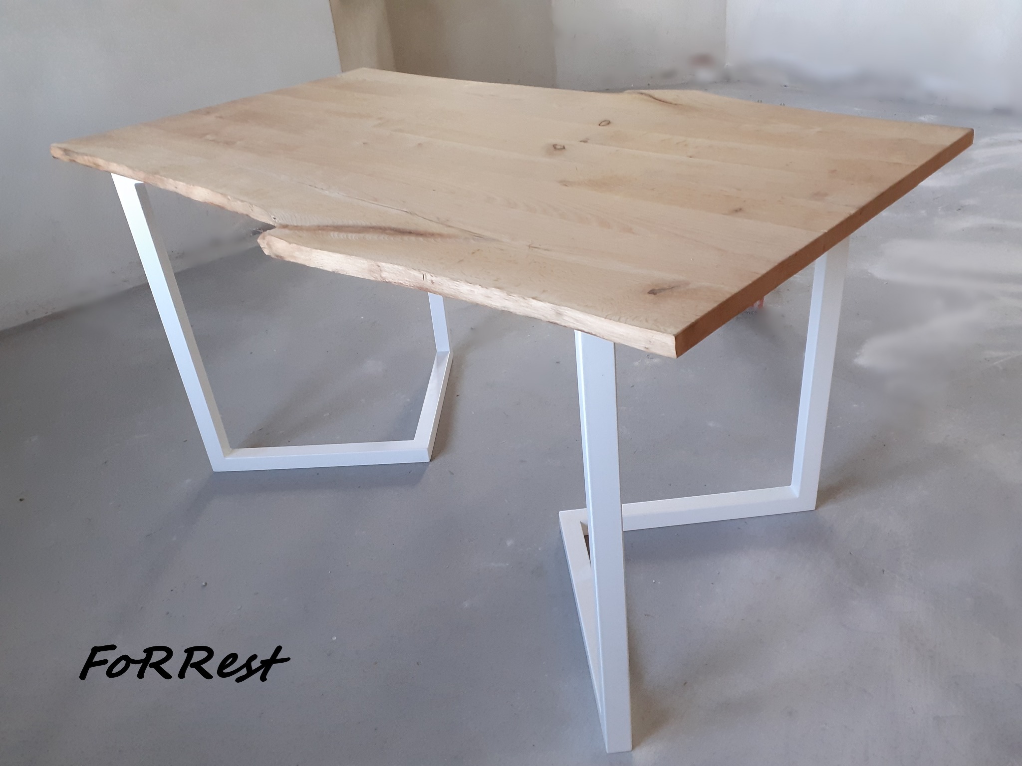 Surowy blat stołu z jasnego drewna o nieregularnym kształcie krawędzi, wsparty na białych, geometrycznych nogach z metalowego profilu, na surowym betonowym podłożu z widocznym logo firmy 'FoRRest'.