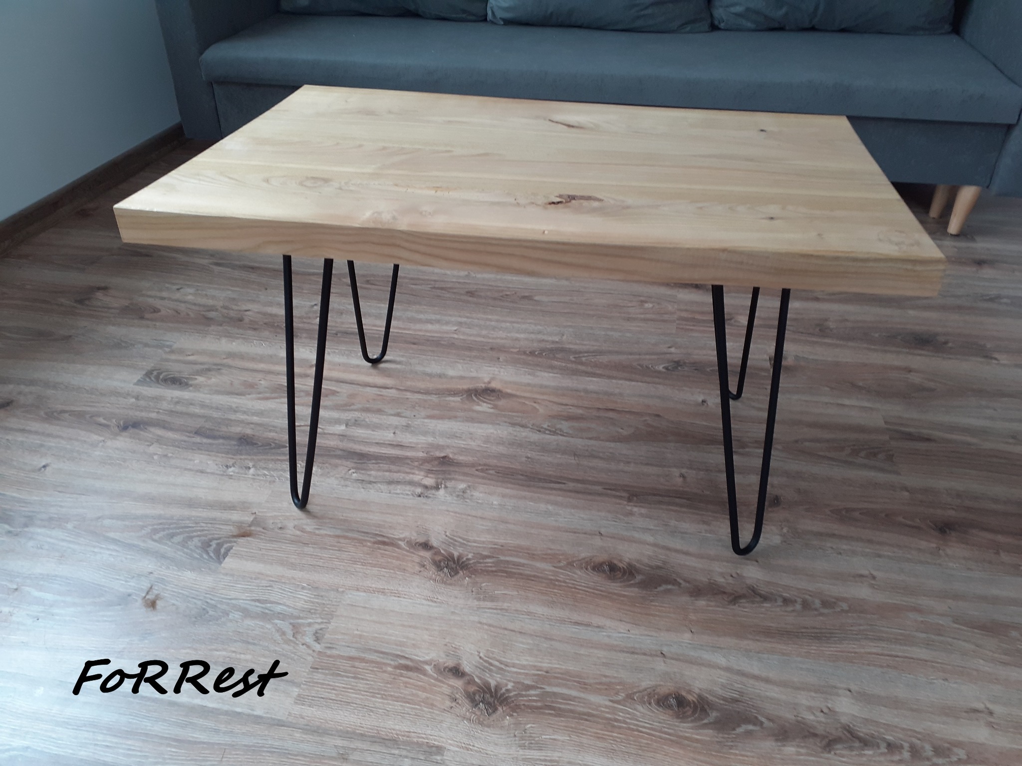 Prostokątny stolik kawowy z blatem z jasnego drewna i czarnymi, metalowymi nogami typu hairpin, ustawiony na panelach podłogowych imitujących drewno, z szarą sofą w tle.