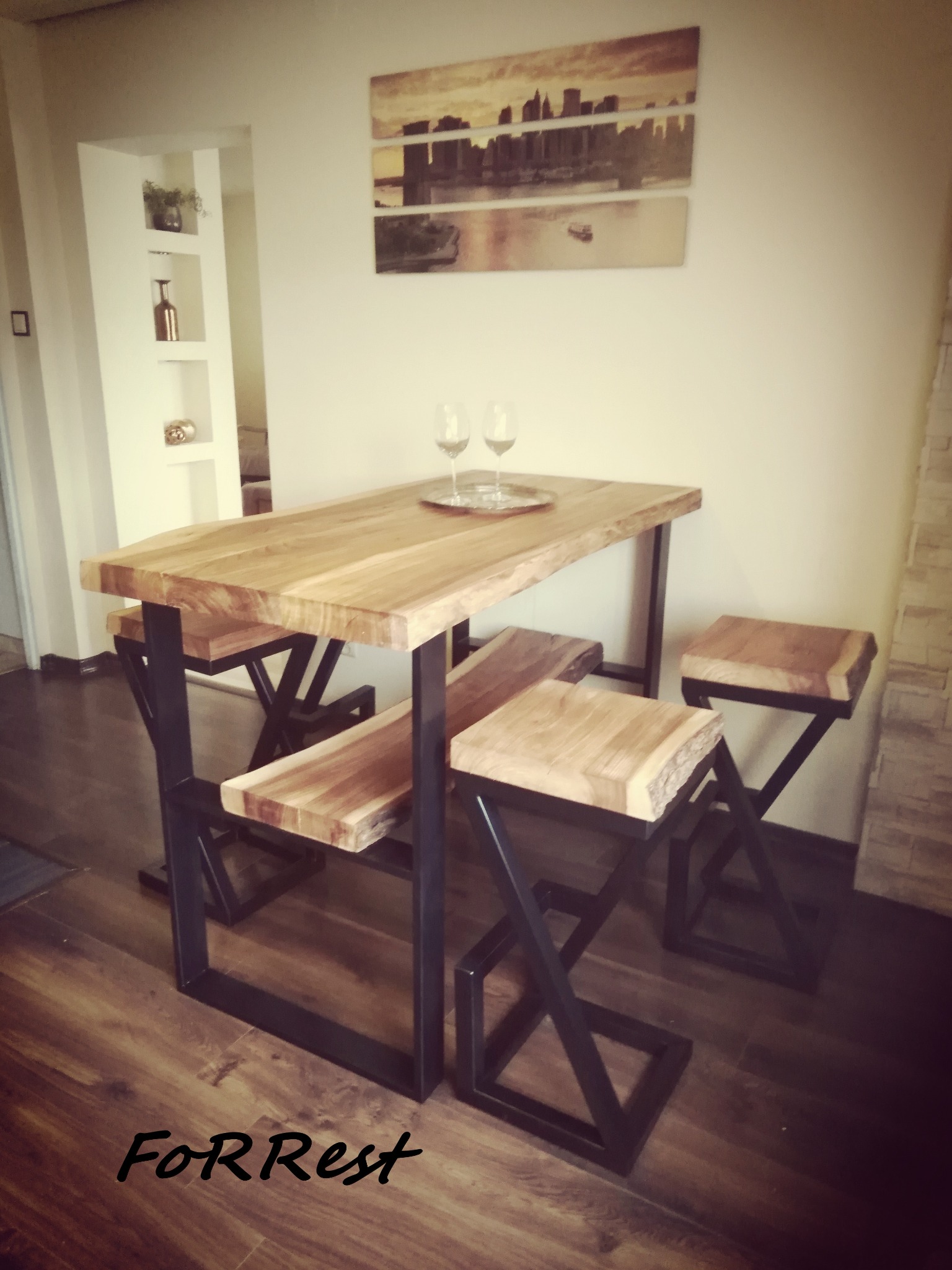 Wysoki stół barowy z blatem z jasnego drewna o nieregularnych krawędziach i metalową podstawą w kształcie litery X, do kompletu drewniane stołki barowe z metalowymi nogami, w tle obraz z motywem...