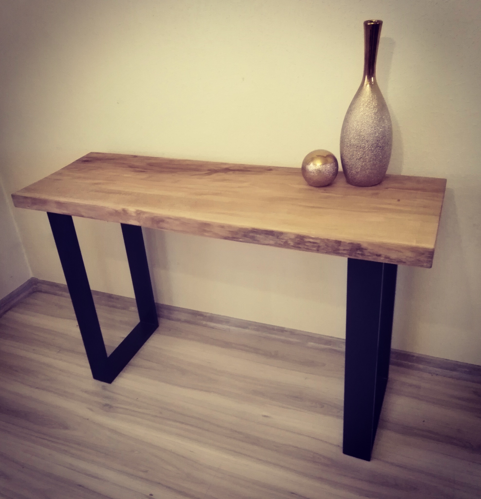 Konsola z blatem z litego drewna o nieregularnych krawędziach i czarnych, metalowych nogach w kształcie litery V, na blacie złota kula i wysoki wazon w odcieniach złota i srebra