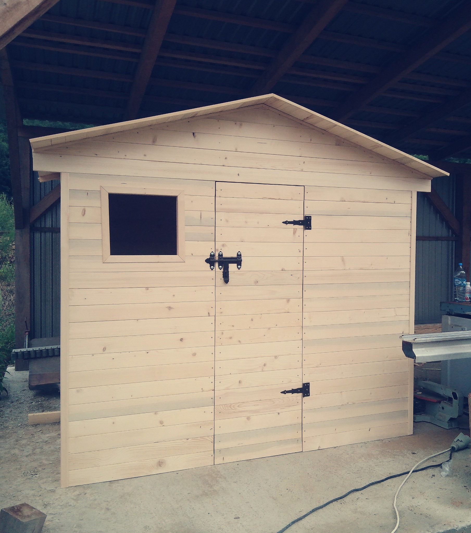 Niewykończona, surowa, drewniana budka narzędziowa z małym oknem i prostymi, czarnymi zawiasami, widoczna w warsztacie obok piły stołowej.