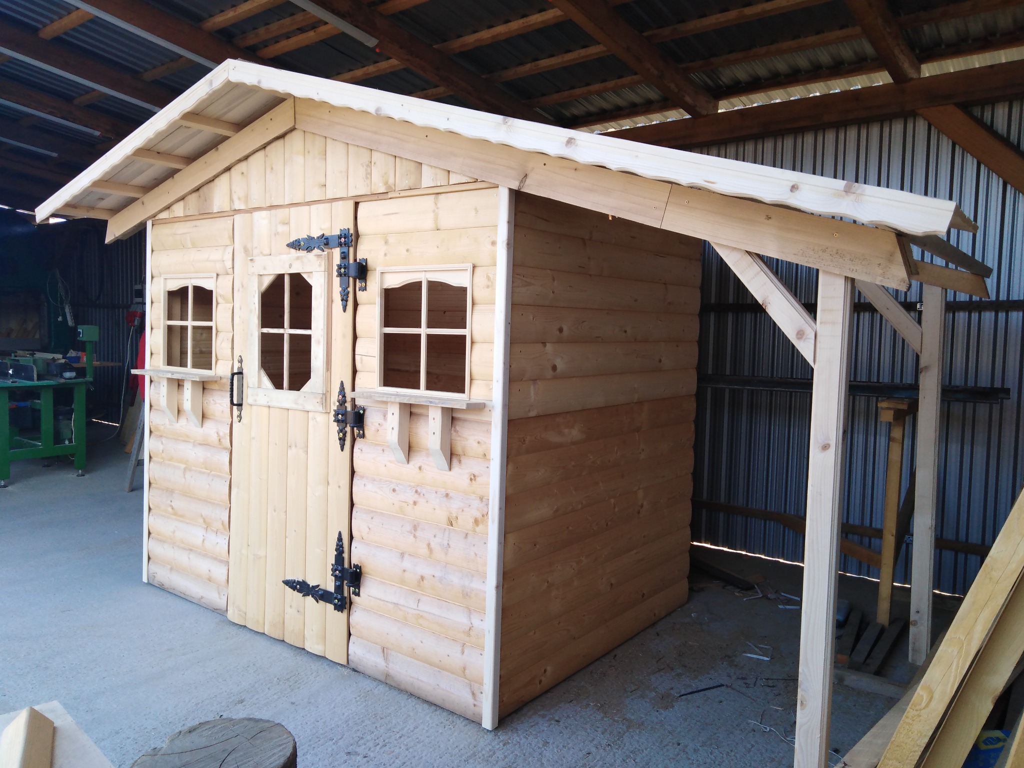 Drewniany domek ogrodowy z dwuspadowym dachem, małymi okienkami z ozdobnymi okiennicami i kutymi zawiasami, częściowo zadaszony, w warsztacie.