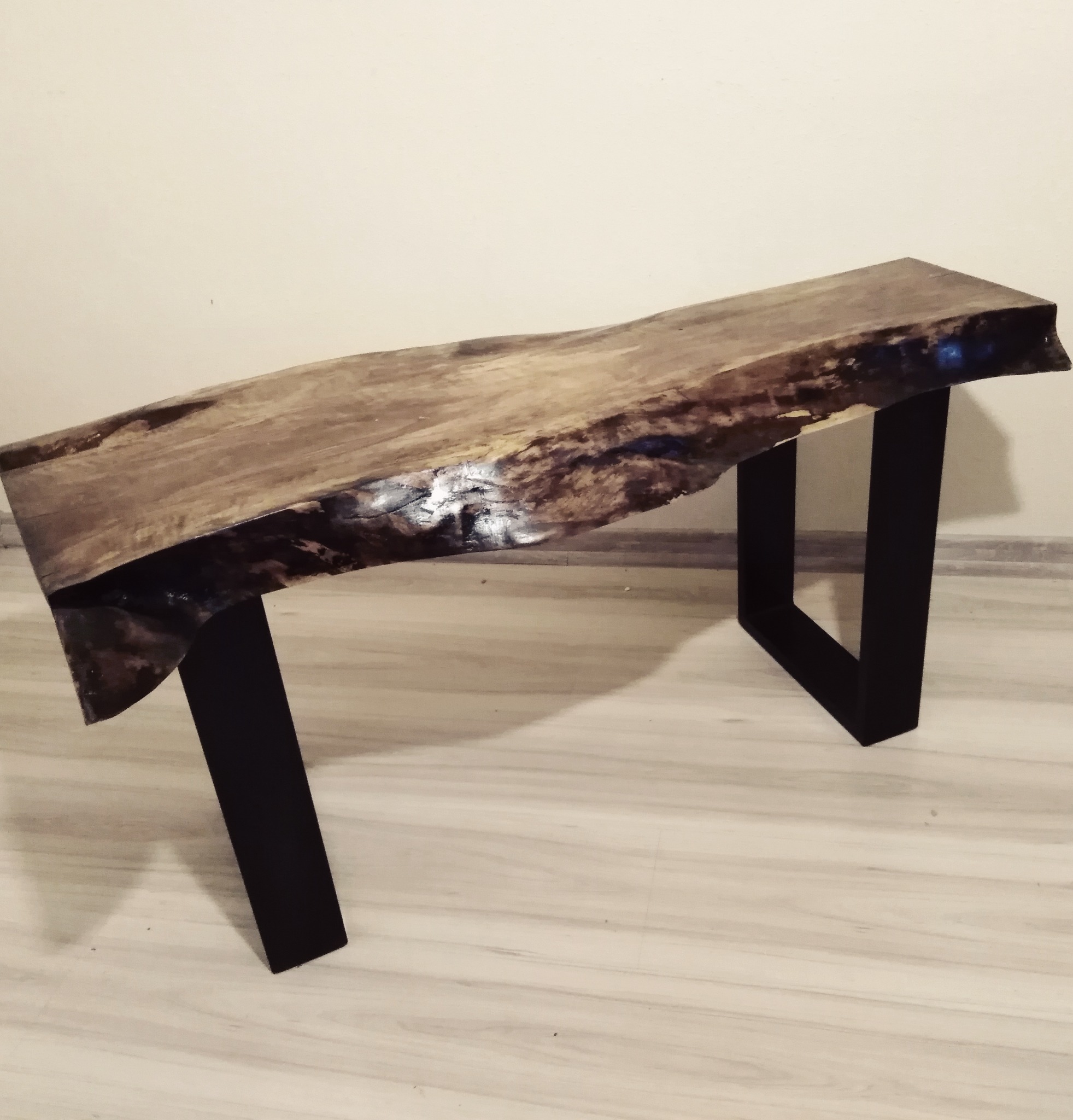 Niski stolik kawowy z blatem z surowego drewna o nieregularnych krawędziach i czarnymi, metalowymi nogami w kształcie trapezu, ustawiony na jasnej podłodze.