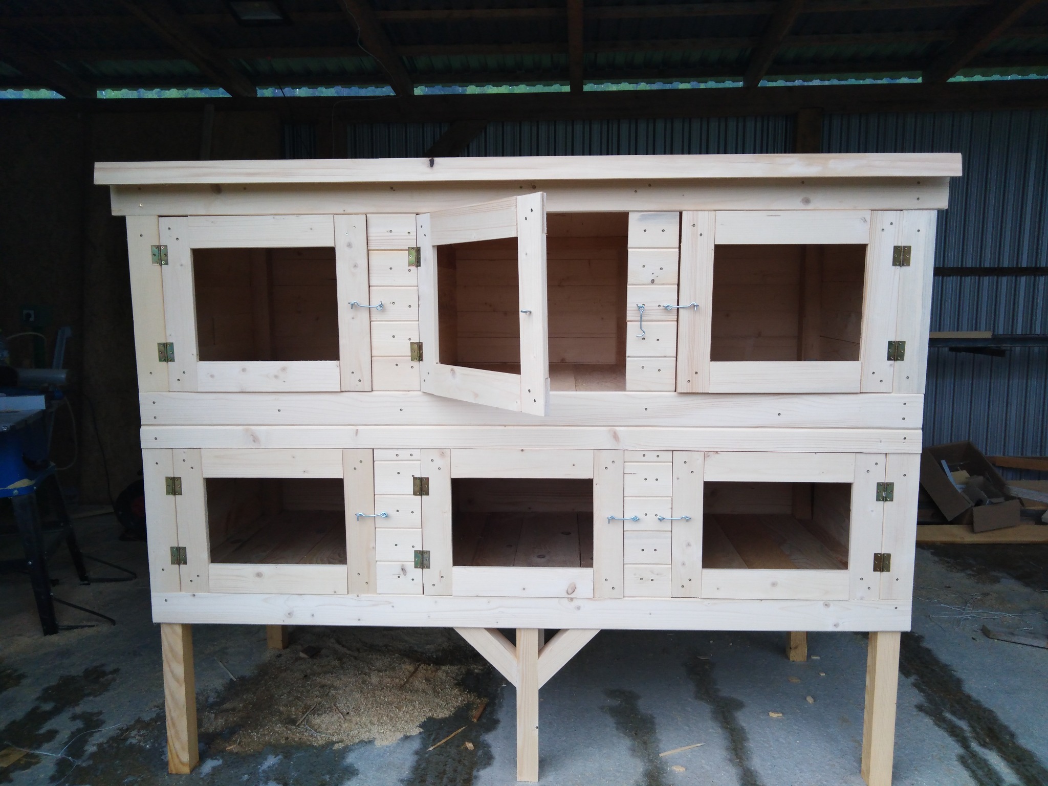 Dwupoziomowa konstrukcja z surowego drewna z sześcioma przegrodami, jedna z drzwiczek otwartych, widoczne zawiasy i haczyki zabezpieczające, całość na czterech nogach z dodatkowym wspornikiem.