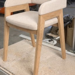 Karol Wincenciak - Beżowe krzesło z drewnianą ramą, tapicerowane siedzisko i oparcie z podłokietnikami, ustawione na dywanie w warsztacie. Widoczne narzędzia stolarskie w tle.