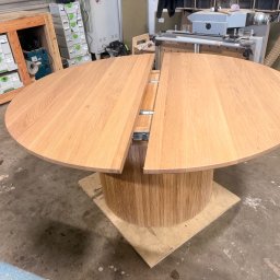 Karol Wincenciak - Okrągły, rozkładany stół z jasnego drewna dębowego na cylindrycznej podstawie, z widocznymi mechanizmami rozsuwania blatu, w warsztacie stolarskim.