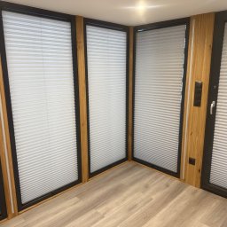 Karol Wincenciak - Nowoczesne okna z plisowanymi roletami w drewnianej obudowie, widok z wnętrza pomieszczenia, jasna podłoga. Minimalistyczny design, równomierne oświetlenie.