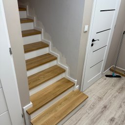 Karol Wincenciak - Jasne, drewniane schody z białymi podstopnicami w korytarzu z panelami podłogowymi i białą, nowoczesną  drzwią. Ściana w odcieniach beżu. Widoczny fragment mopa.