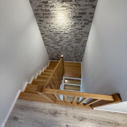 Karol Wincenciak - Drewniane schody z balustradą, widok z góry, z fragmentem buta w rogu. Ściana wykończona dekoracyjnym kamieniem. Jasne panele podłogowe.