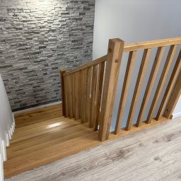 Karol Wincenciak - Jasne, drewniane schody z balustradą, widok z góry. Ściana wykończona szarą imitacją kamienia, podłoga z paneli. Nowoczesne wykończenie wnętrza.