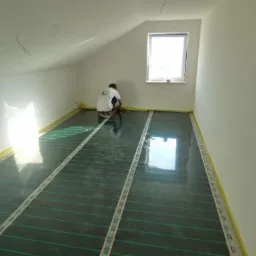Instalacja mat grzewczych na podłodze w pokoju na poddaszu, widoczny pracownik w białej koszulce przyklejający taśmę mocującą do podłoża, okno w tle.