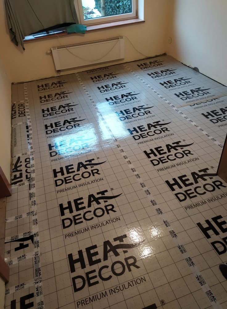 Przygotowanie podłoża pod ogrzewanie podłogowe: rozłożone płyty izolacyjne Heat Decor z nadrukowanym logo na podłodze w pomieszczeniu z grzejnikiem pod oknem.