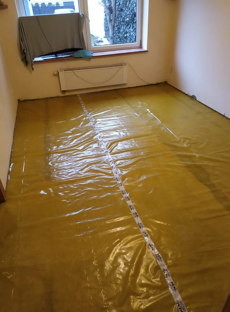 Przygotowanie do montażu ogrzewania podłogowego: żółta folia izolacyjna rozłożona na podłodze z taśmą mierniczą HEAT DECOR, widoczny grzejnik pod oknem.