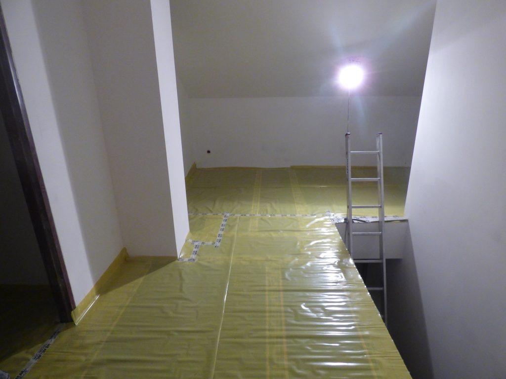 Przygotowanie do montażu ogrzewania podłogowego: żółta folia izolacyjna rozłożona na podłodze, drabina aluminiowa ustawiona w pomieszczeniu o białych ścianach.