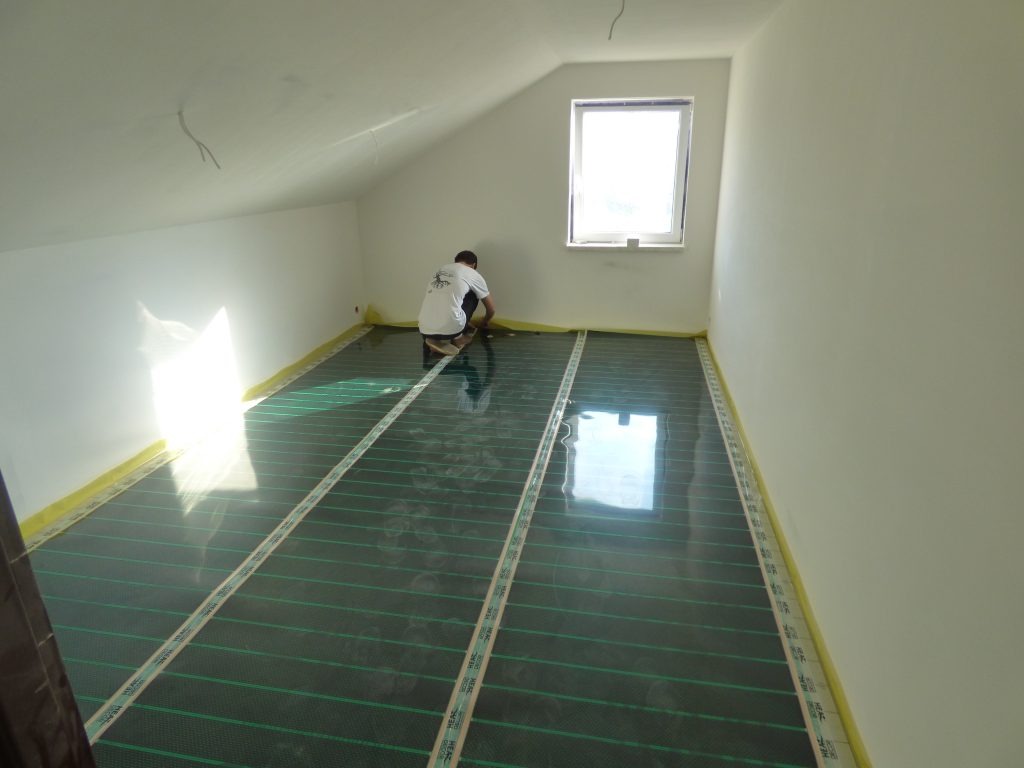 Instalacja mat grzewczych na podłodze w pokoju na poddaszu, widoczny pracownik w białej koszulce przyklejający taśmę mocującą do podłoża, okno w tle.