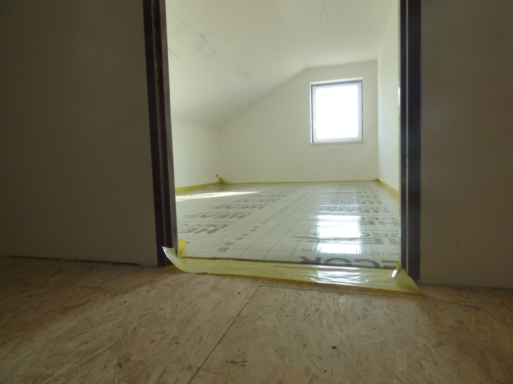 Przygotowanie do montażu ogrzewania podłogowego: rozłożona mata izolacyjna z folią aluminiową na podłodze w pomieszczeniu ze skosami i oknem, widok zza progu.