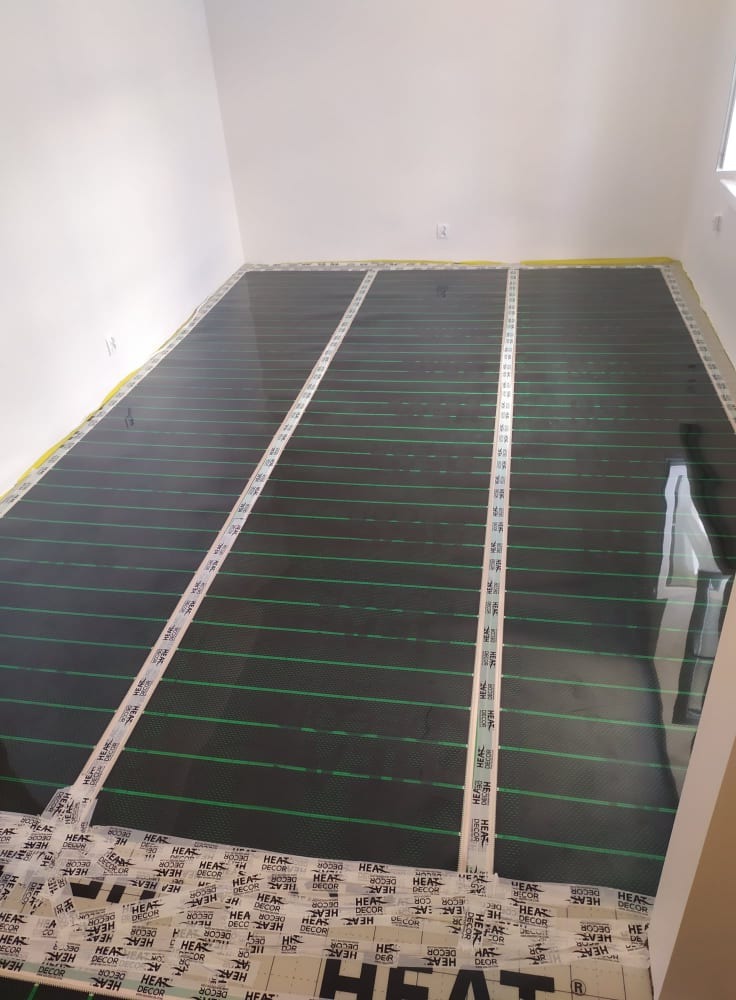Układanie mat grzewczych pod ogrzewanie podłogowe w pomieszczeniu w Konstantynowie Łódzkim, widoczne taśmy mocujące i zielone linie grzewcze na czarnej macie.