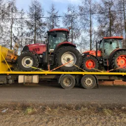 Żółta ciężarówka transportuje dwa traktory: czerwony Case IH Puma i pomarańczowy Kubota M8560, zabezpieczone pasami transportowymi na platformie. W tle widoczne nagie drzewa i błękitne niebo.
