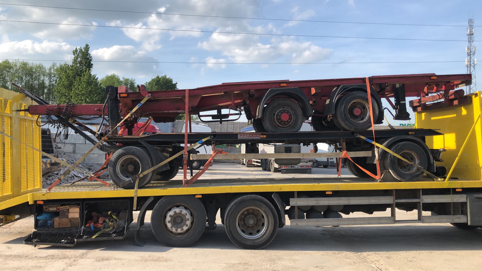 Żółta laweta przewozi czerwoną naczepę, zabezpieczoną pasami transportowymi, widoczne skrzynki narzędziowe i elementy mocujące. W tle drzewa i słup telekomunikacyjny.