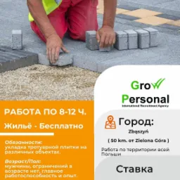 Pracownik w odblaskowej kamizelce układa szarą kostkę brukową na przygotowanym podłożu z grysu, widoczny fragment ułożonej powierzchni z kostki w różnych odcieniach szarości i beżu.