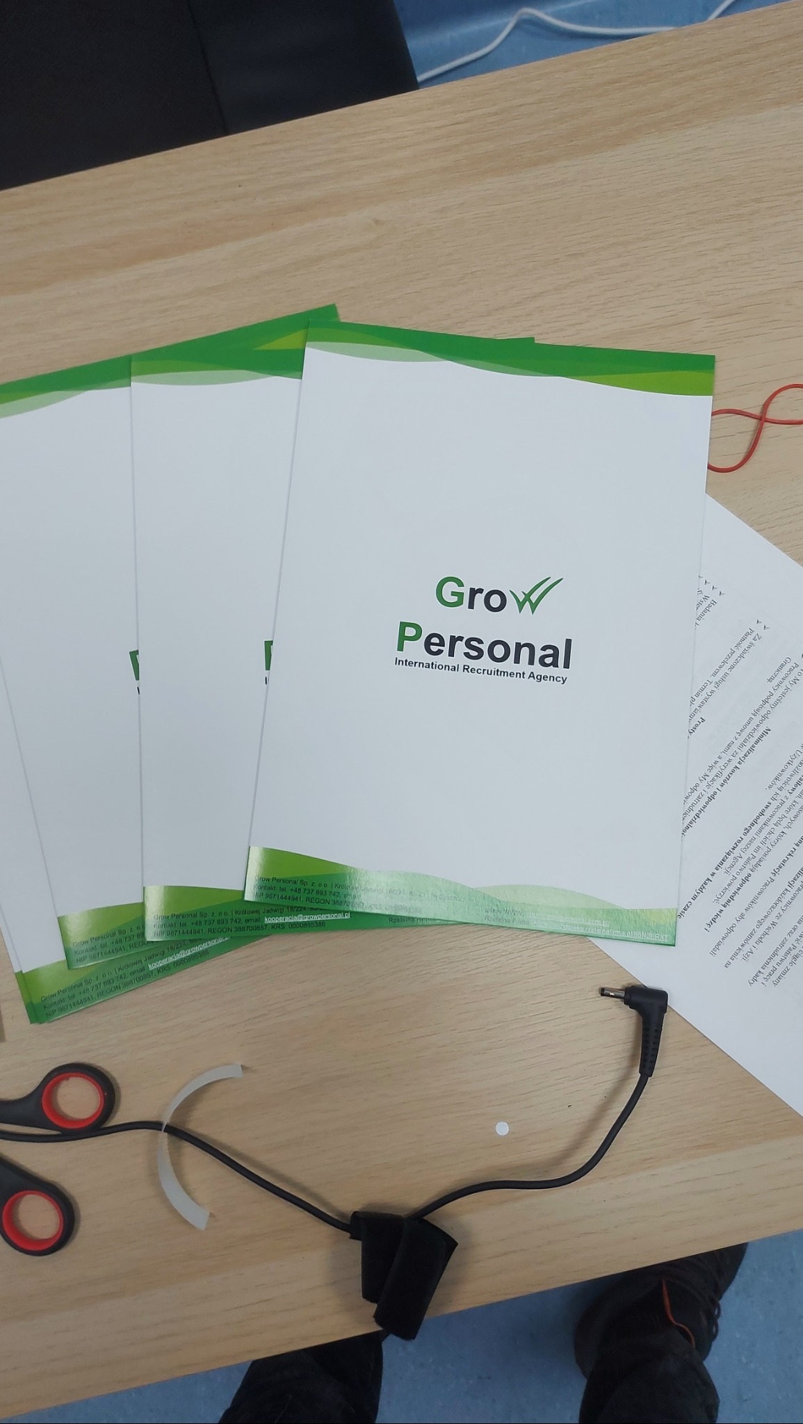 Stos ulotek firmy rekrutacyjnej Grow Personal na drewnianym biurku, obok nożyczki z czerwonymi uchwytami i czarny kabel z zasilaczem.