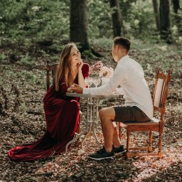 Aleja Fotografii - Para siedząca przy stoliku w lesie, kobieta w długiej czerwonej sukni, mężczyzna w białej koszuli i krótkich spodenkach, na stoliku kwiaty i kieliszki.