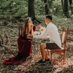 Para siedząca przy stoliku w lesie, kobieta w długiej czerwonej sukni, mężczyzna w białej koszuli i krótkich spodenkach, na stoliku kwiaty i kieliszki.