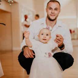 Aleja Fotografii - Uśmiechnięty tata przykuca obok małej dziewczynki w białej sukience z kwiatami, trzymając ją za rękę w jasnym wnętrzu.