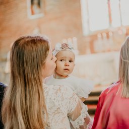 Aleja Fotografii - Matka całuje w policzek niemowlę z opaską w kwiaty na głowie, w tle wnętrze kościoła z oknami witrażowymi i świecami.