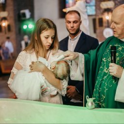 Aleja Fotografii - Ksiądz w zielonej szacie udziela chrztu dziecku trzymanemu przez matkę w kościele, ojciec obserwuje w tle
