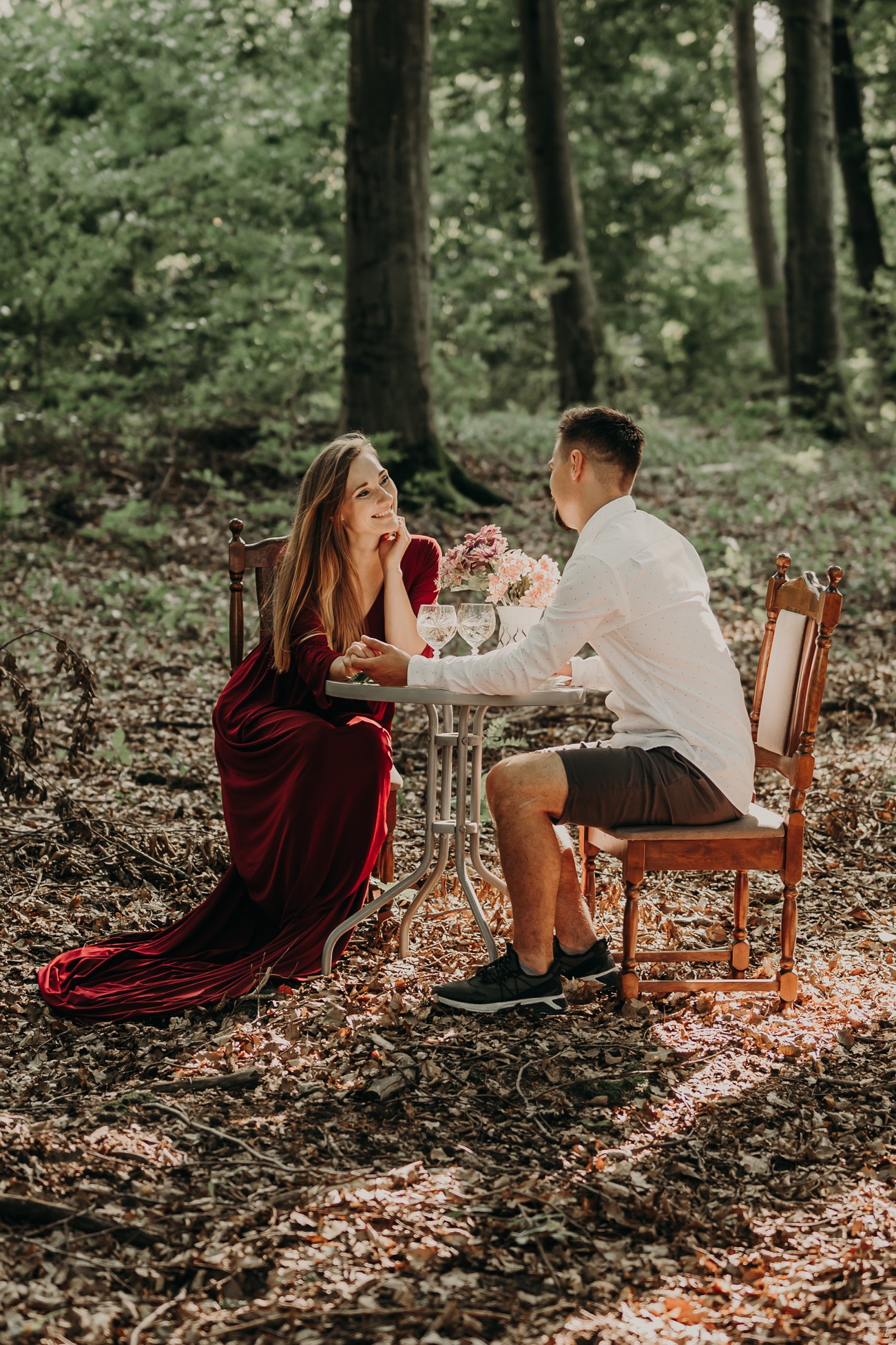 Para siedząca przy stoliku w lesie, kobieta w długiej czerwonej sukni, mężczyzna w białej koszuli i krótkich spodenkach, na stoliku kwiaty i kieliszki.
