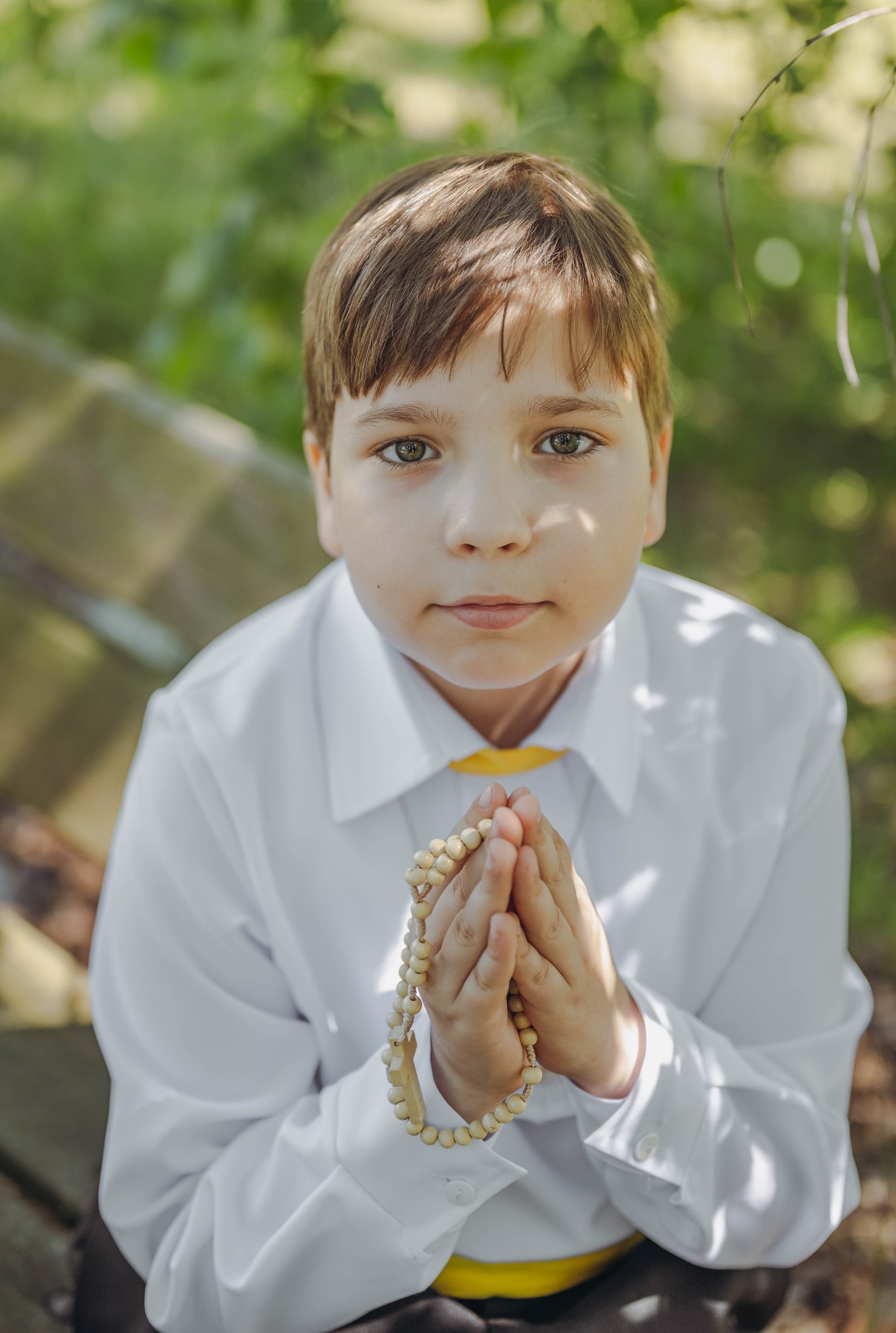 Chłopiec w białej koszuli z żółtą muchą, trzymający w dłoniach drewniany różaniec, uchwycony na sesji w Lubawie, z delikatnym, naturalnym oświetleniem.