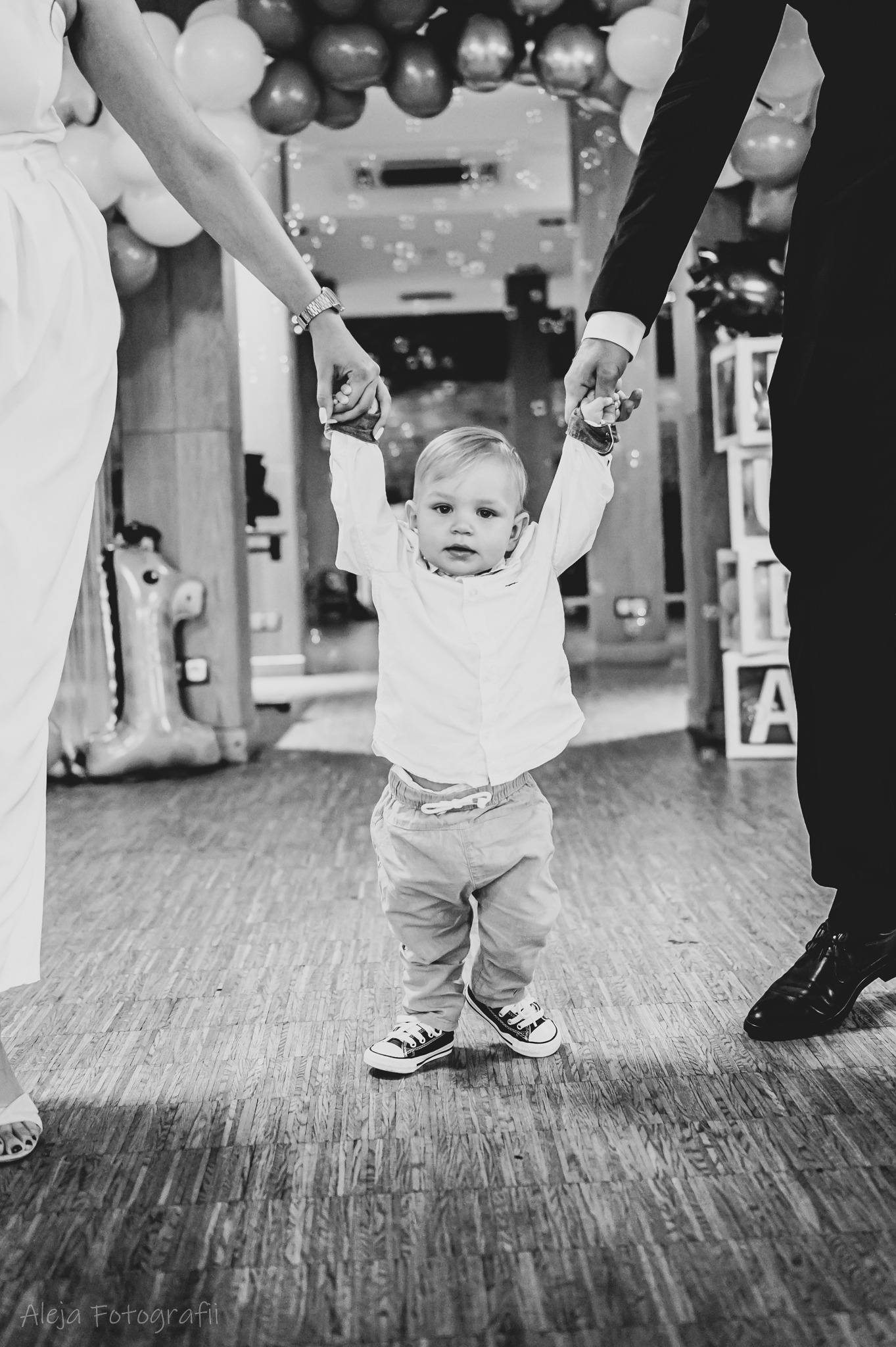 Czarno-białe zdjęcie małego chłopca trzymanego za ręce przez rodziców, ubrany w koszulę i spodnie, z balonami i bańkami mydlanymi w tle.