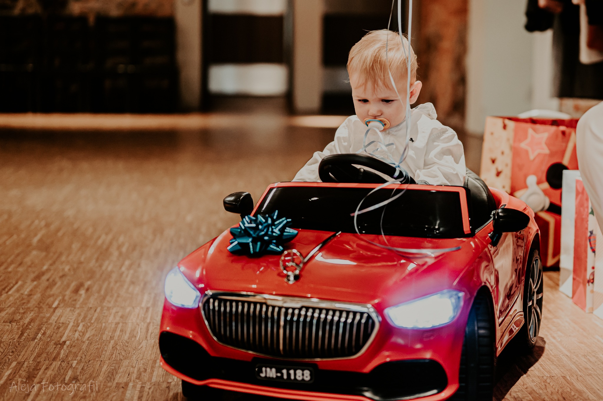 Mały chłopiec z blond włosami siedzi w czerwonym, elektrycznym samochodzie dla dzieci z niebieską kokardą na masce, trzymając w buzi smoczek i otoczony wstążkami.