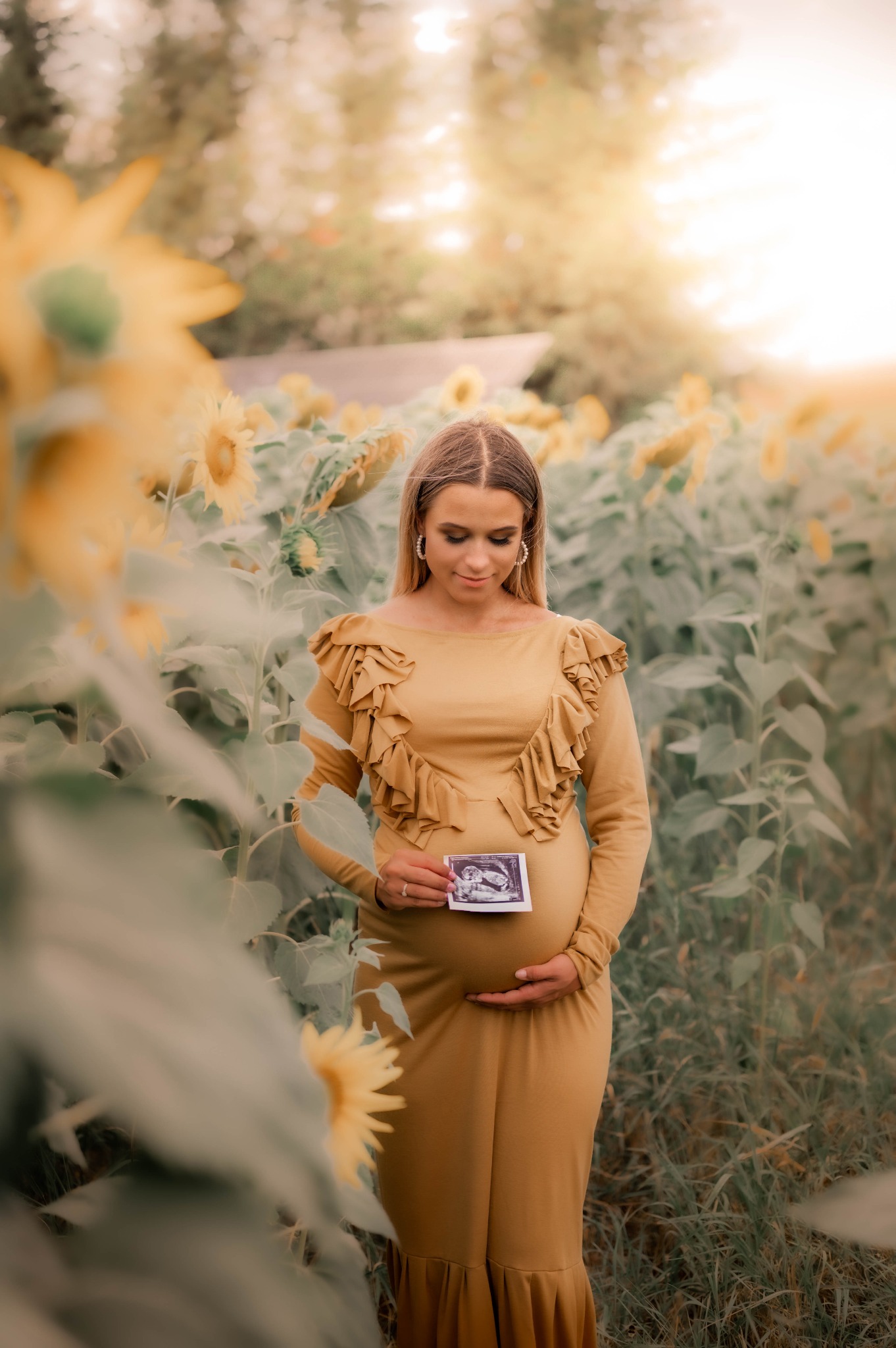 Kobieta w złotej sukni, otoczona polem słoneczników, trzyma zdjęcie USG na brzuchu.