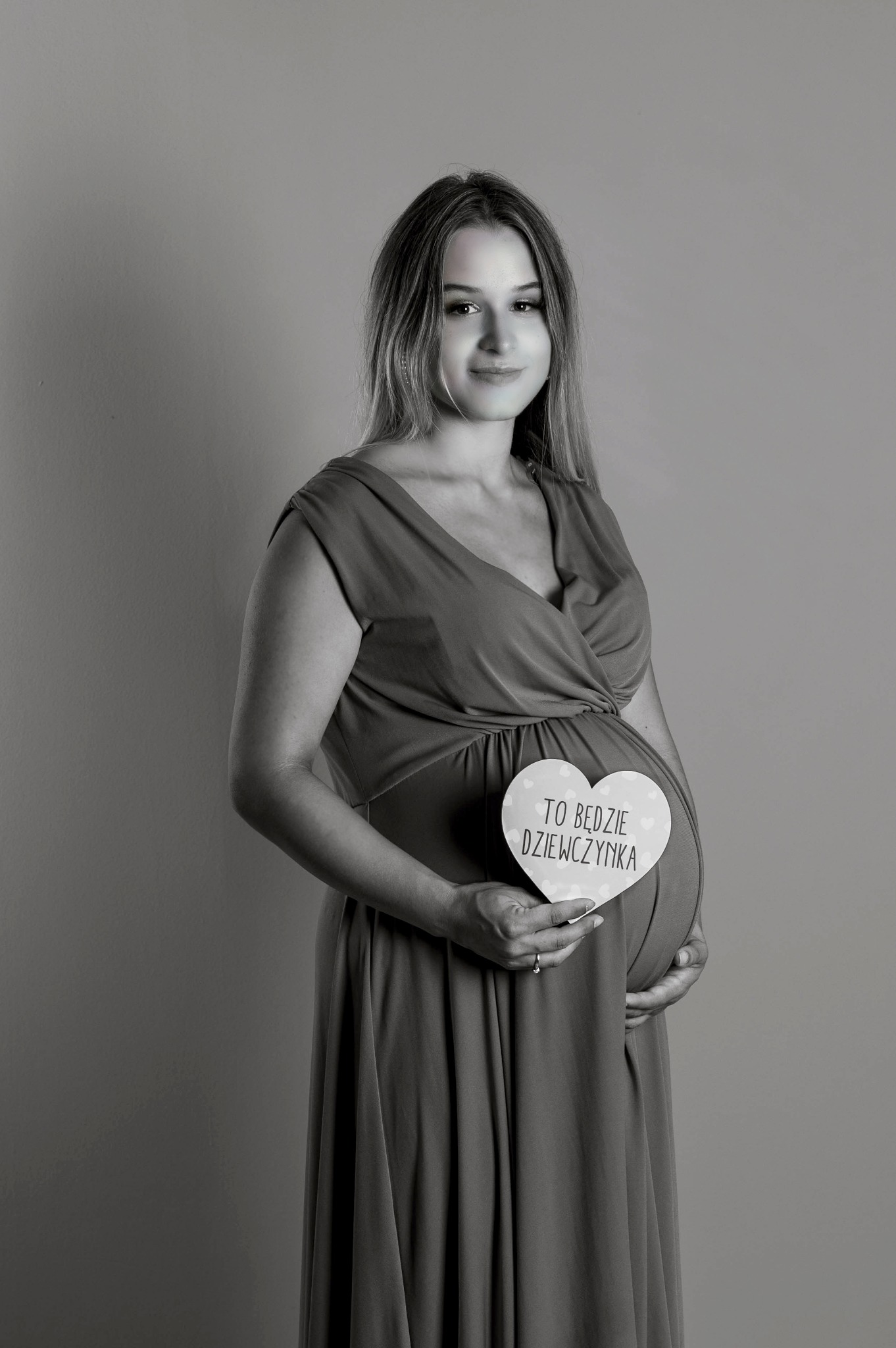 Czarno-białe zdjęcie uśmiechniętej kobiety w sukience ciążowej trzymającej serce z napisem 'To będzie dziewczynka' na tle jednolitej ściany.