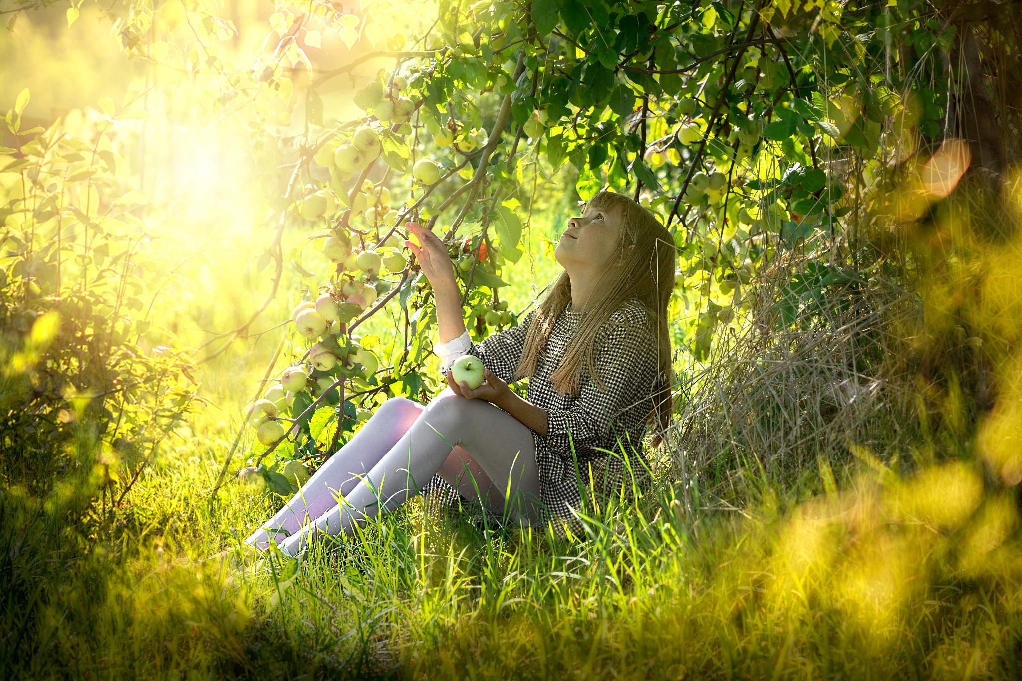 Młoda kobieta w kwiecistej bluzce i legginsach siedzi pod jabłonią, trzyma zielone jabłko i wyciąga dłoń w kierunku owoców, w tle jasne światło słoneczne i bujna zieleń.
