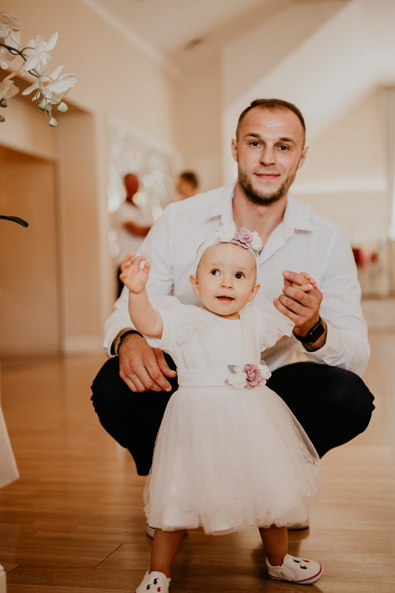 Uśmiechnięty tata przykuca obok małej dziewczynki w białej sukience z kwiatami, trzymając ją za rękę w jasnym wnętrzu.