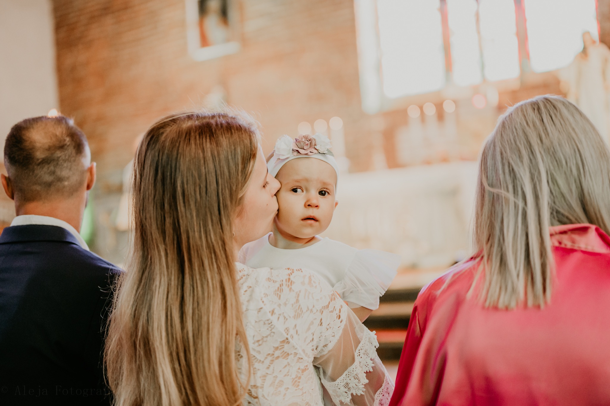 Matka całuje w policzek niemowlę z opaską w kwiaty na głowie, w tle wnętrze kościoła z oknami witrażowymi i świecami.