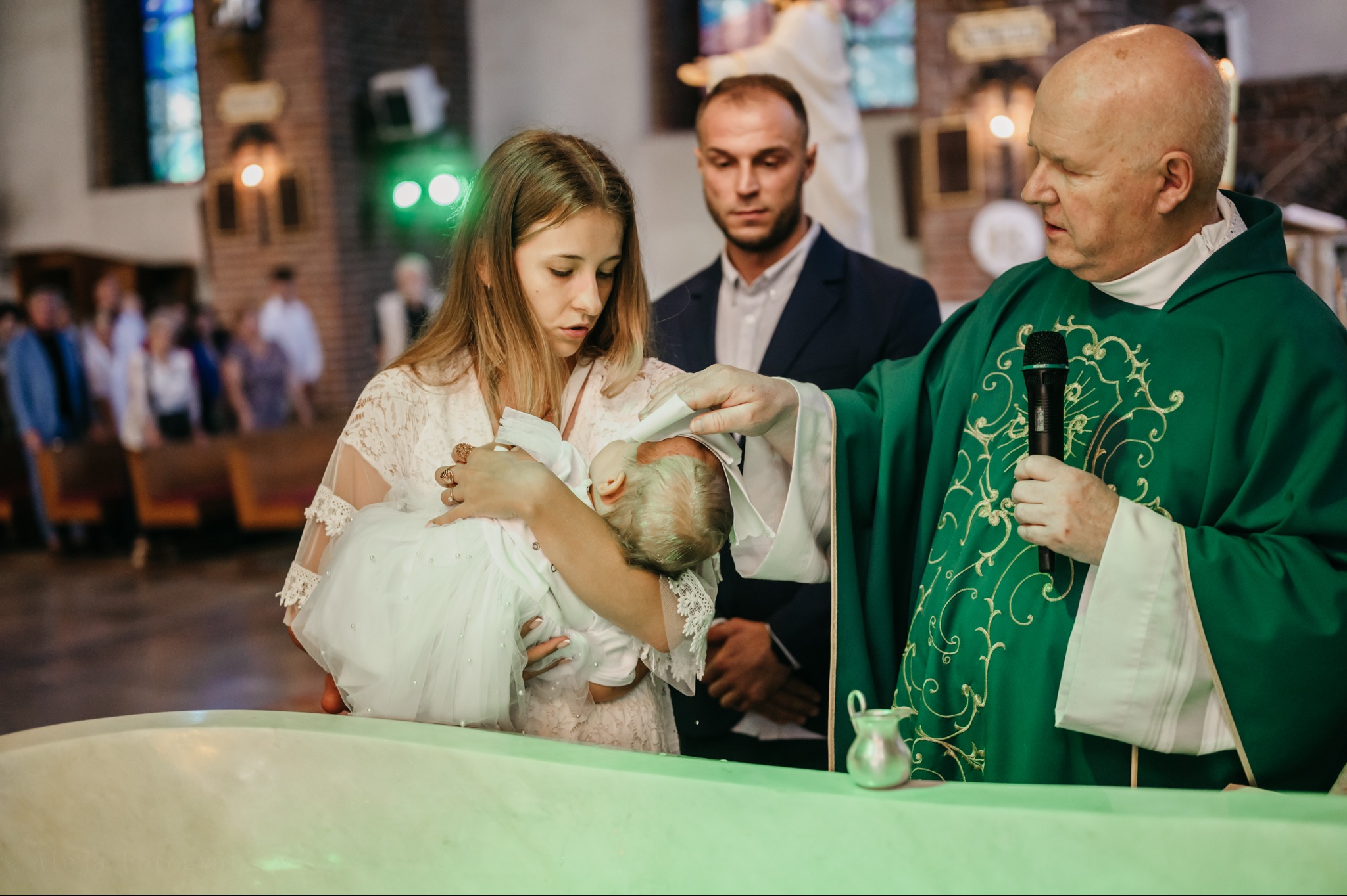 Ksiądz w zielonej szacie udziela chrztu dziecku trzymanemu przez matkę w kościele, ojciec obserwuje w tle