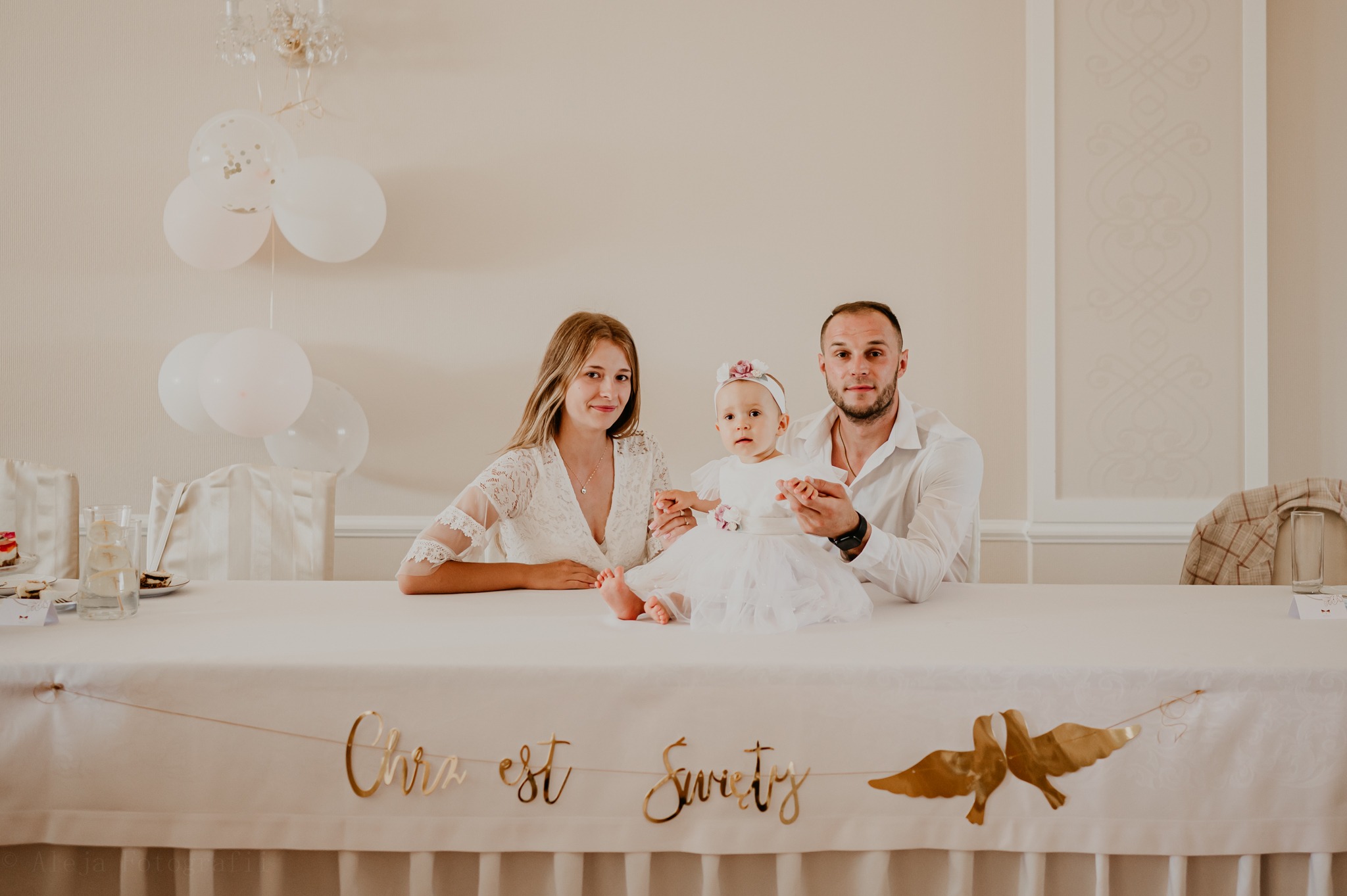 Rodzina pozuje za stołem z napisem 'Chrzest Święty' i złotymi gołębiami, białe balony w tle.
