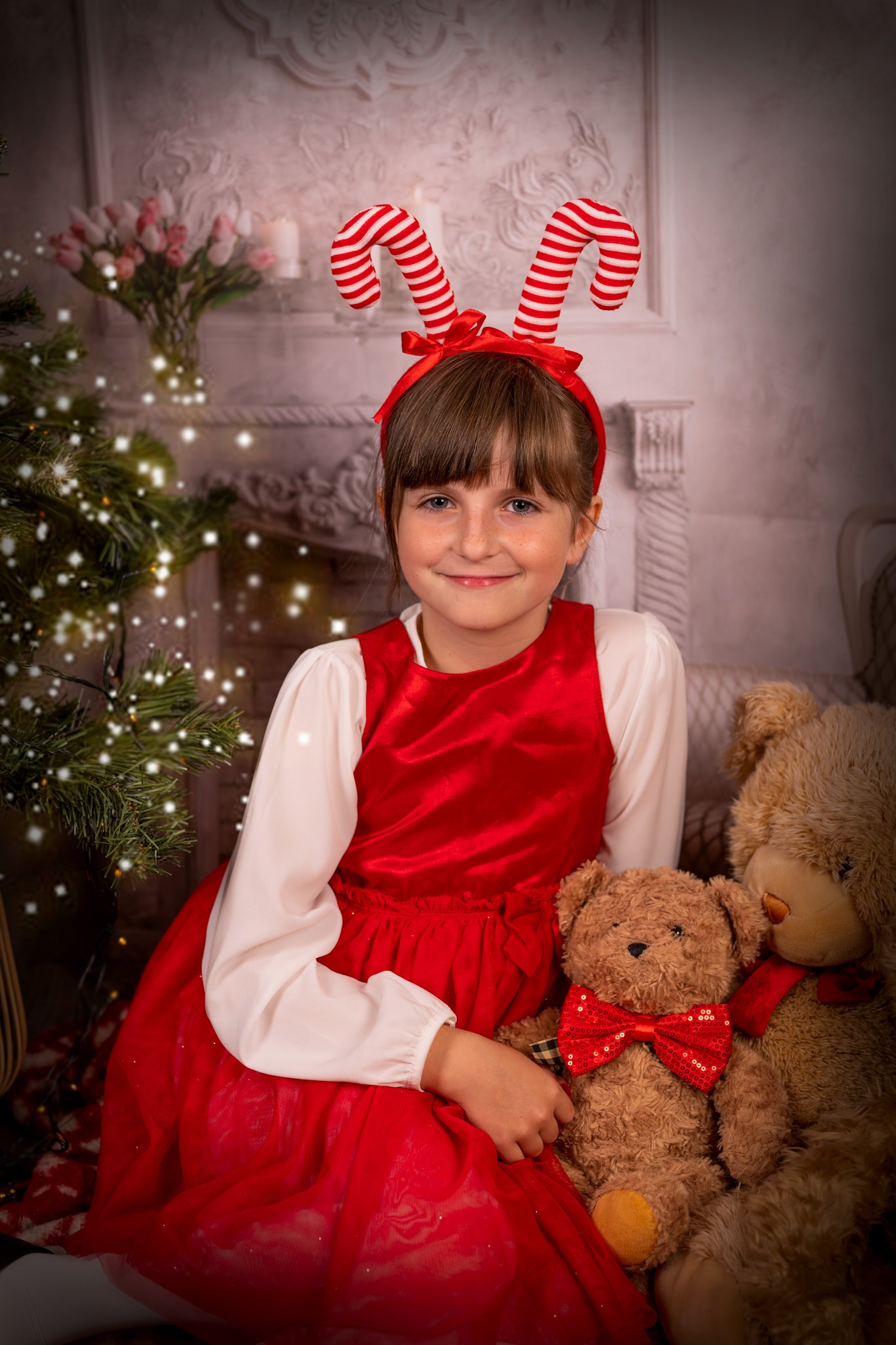 Uśmiechnięta dziewczynka w czerwonej sukience i opasce z lizakami, siedząca z pluszowymi misiami na tle choinki i kominka.