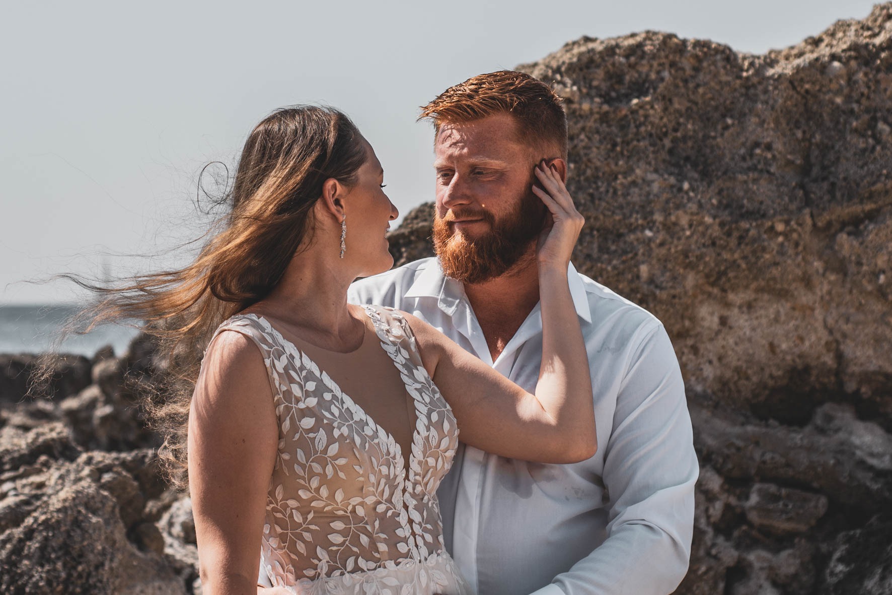 Zakochana para: kobieta w sukni ślubnej z koronkowymi aplikacjami dotyka twarzy rudowłosego mężczyzny z brodą na tle skalistego wybrzeża w słoneczny dzień.