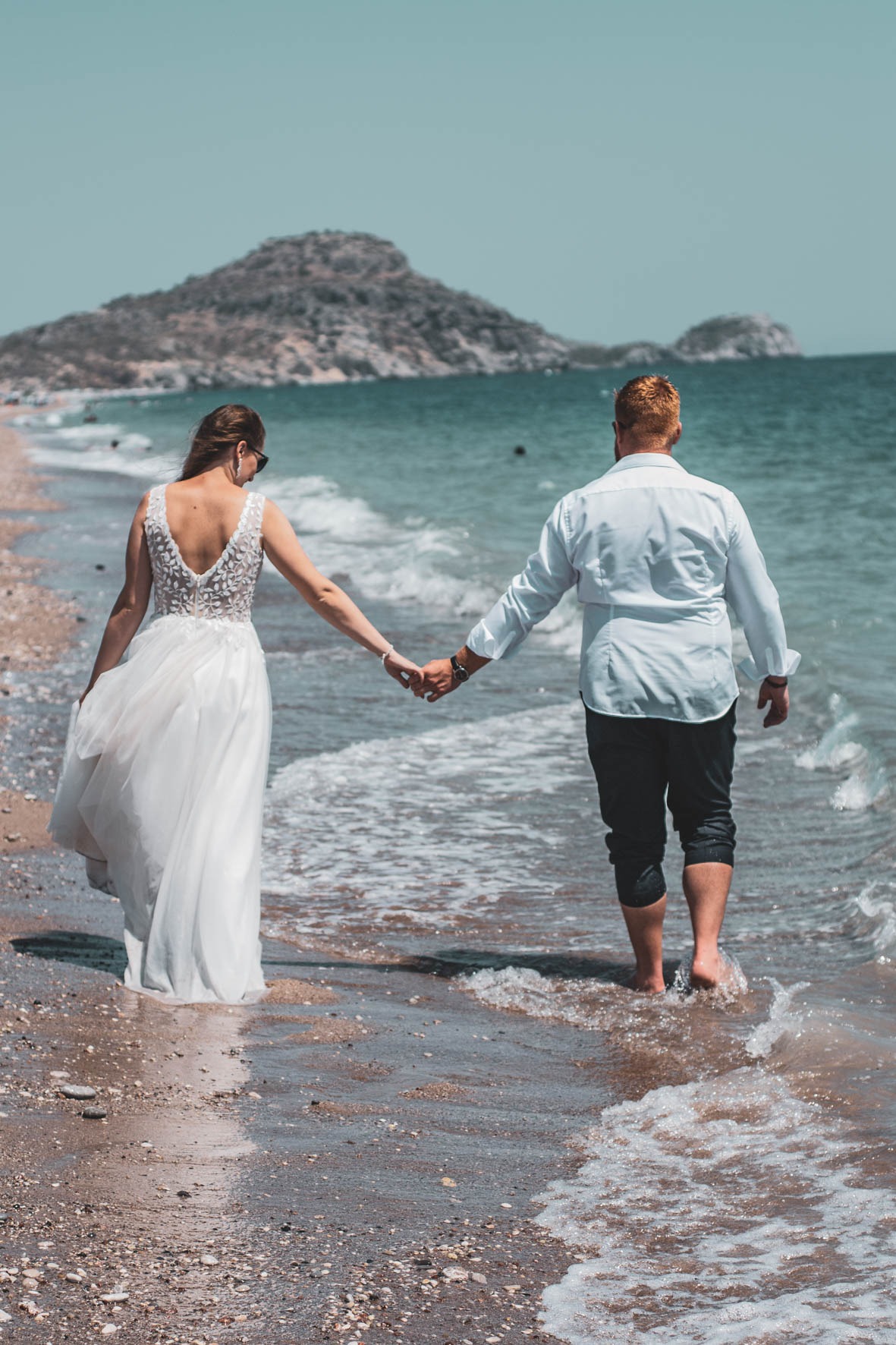 Para młoda spaceruje brzegiem morza trzymając się za ręce, widok z tyłu, kobieta w białej sukni z koronkowymi zdobieniami na plecach, mężczyzna w białej koszuli i ciemnych spodniach podwiniętych...