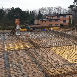 BUD-MAR Marcin Gorzkowski - Zbrojenie płyty fundamentowej z widocznym szalunkiem i pracownikami budowlanymi w tle, na tle budynku mieszkalnego i drzew