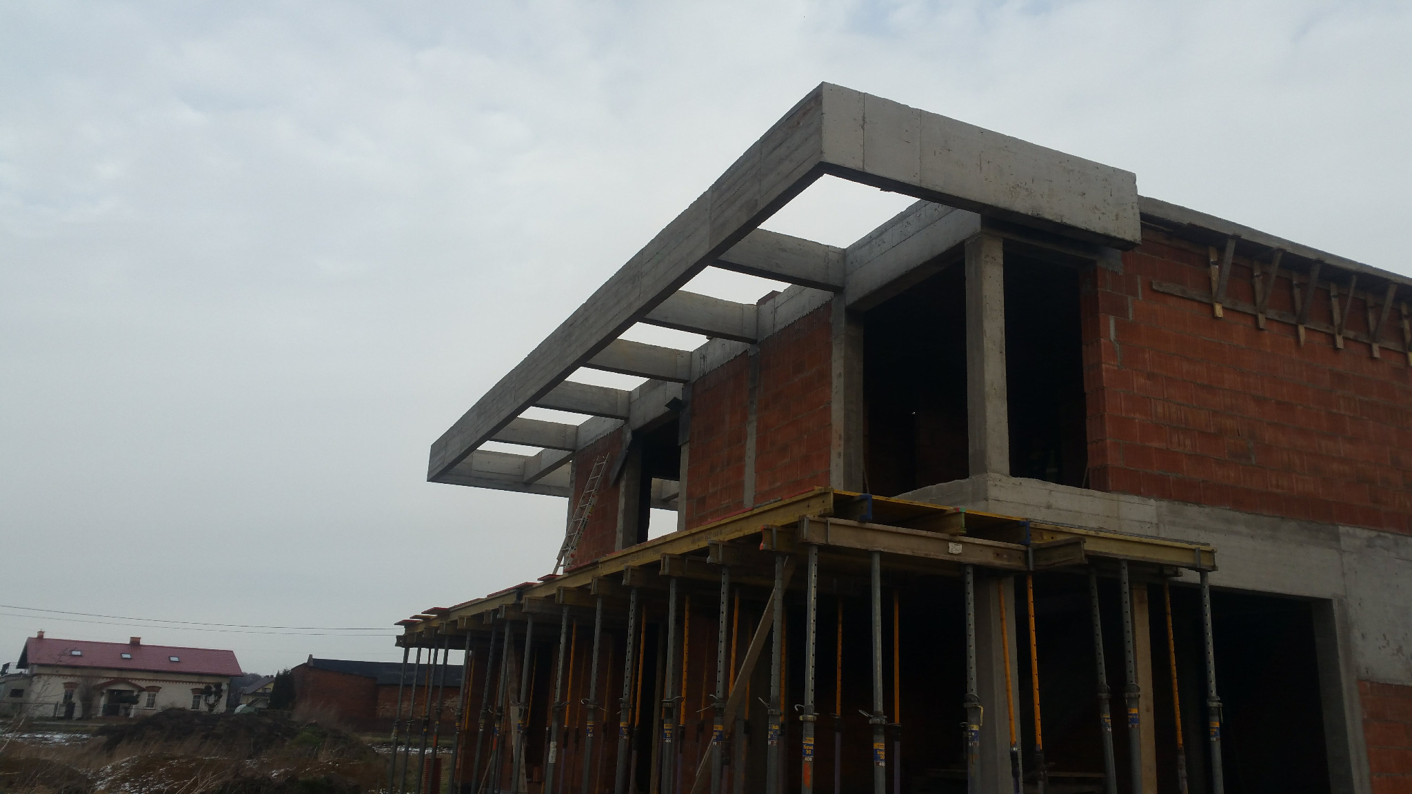Surowy stan domu murowanego z betonu komórkowego, widoczne szalunki i podpory stropu, w tle budynek z czerwoną dachówką.