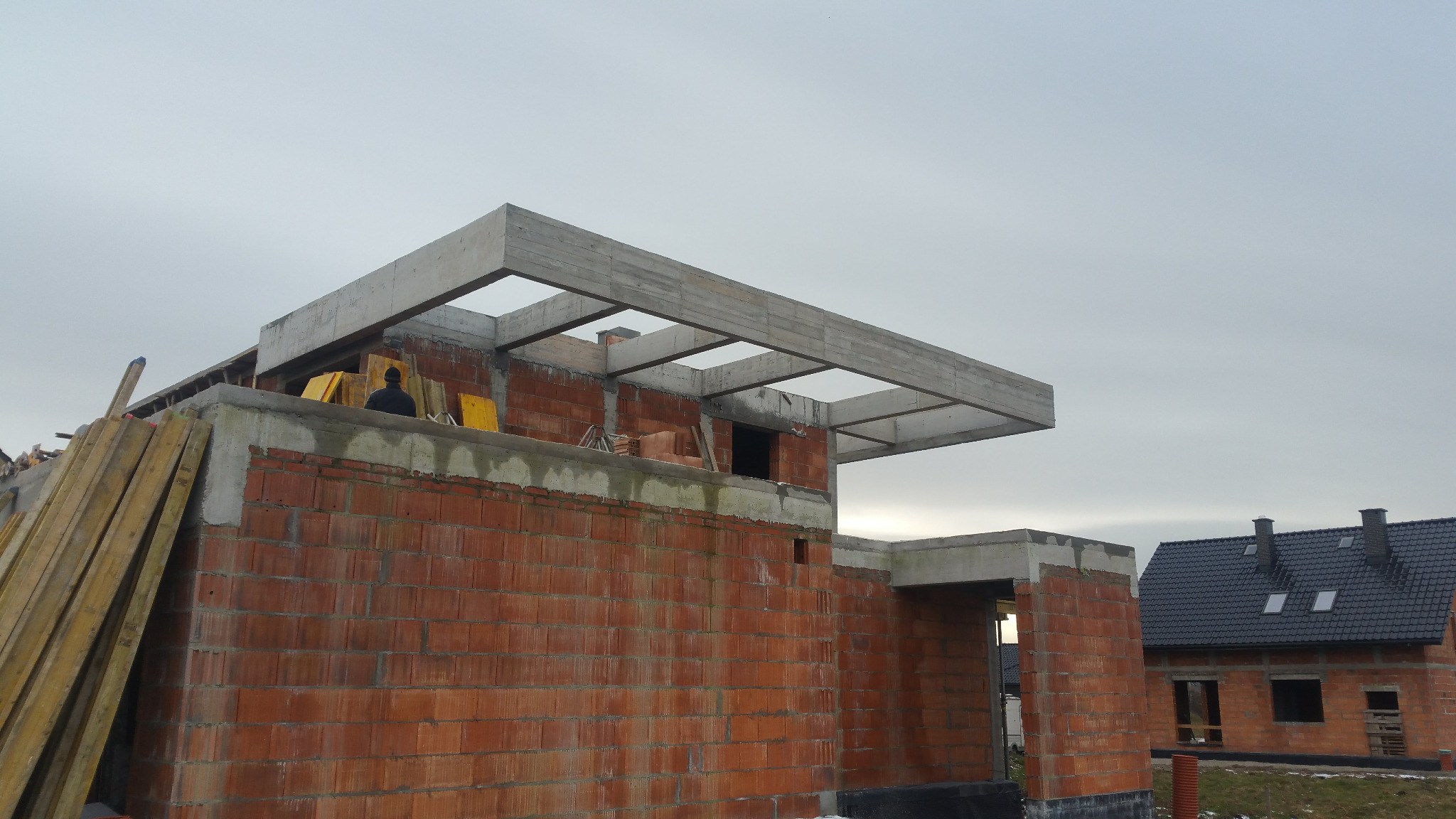 Budowa domu w stanie surowym otwartym: czerwona cegła, betonowe nadproża, widoczne deski szalunkowe, pracownik w czarnej kurtce na tle pochmurnego nieba.