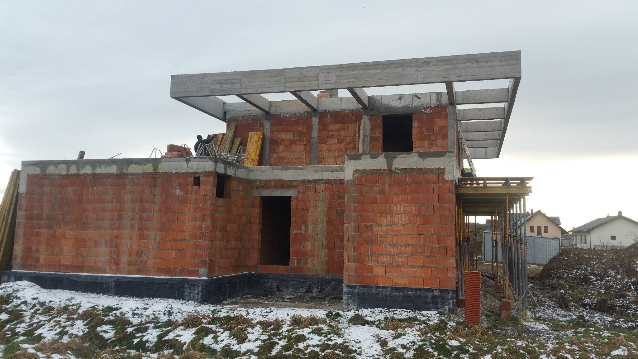 Surowy stan budynku jednorodzinnego w trakcie budowy, widoczne ściany z czerwonej cegły, betonowe nadproża, częściowo wykonana izolacja przeciwwilgociowa fundamentów, w tle krajobraz...