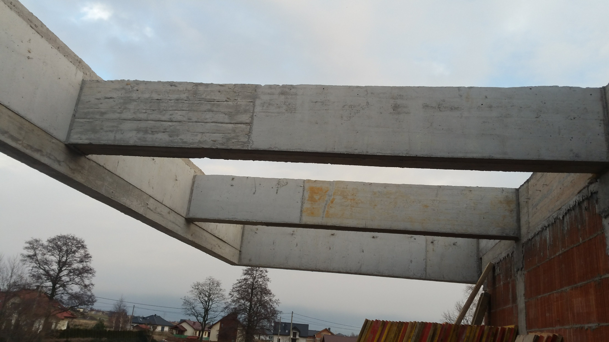 Surowa konstrukcja betonowa stropu w trakcie budowy domu jednorodzinnego, widok z dołu na tle pochmurnego nieba i zabudowań.